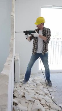 Koruyucu kask ve eldiven takan inşaat işçisi iç duvarı yıkmak için döner çekiç matkabı kullanıyor ve ev yenileme projesi sırasında yerde enkaz yığını oluşturuyor.