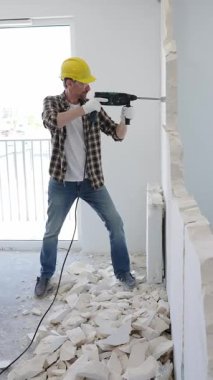 Koruyucu kask ve eldiven takan inşaat işçisi iç duvarı yıkmak için döner çekiç matkabı kullanıyor ve ev yenileme projesi sırasında yerde enkaz yığını oluşturuyor.