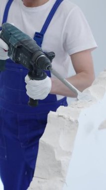 İnşaat işçisi, beyaz duvarı döner çekiç matkabıyla yıkıyor, mavi tulum ve koruyucu beyaz eldivenler giyiyor, yenileme projesi sırasında toz üretiyor, yakın çekim görüntüsü sağlıyor..