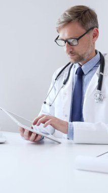 Laboratuvar önlüğü ve steteskop giyen orta yaşlı doktor, tıp bürosunda masasında otururken dijital tablet kullanıyor, ön planda da da tıbbi çizelge ve kalem var. Tıp konsepti.
