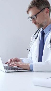 Orta yaşlı doktor özenle bir dizüstü bilgisayarda yazıyor, muhtemelen hasta kayıtlarını güncelliyor ya da araştırma yapıyor, sağlık ve teknolojinin tıbbi ortamdaki kesişimini gösteriyor. İlaç..