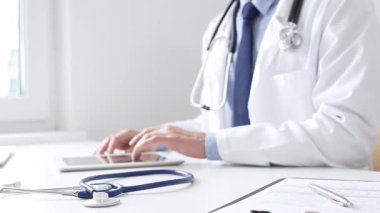 Klinikte dijital tablet kullanan doktor adamın yanındaki masada mavi steteskop duruyor. Tıp ve sağlık hizmetleri kavramı
