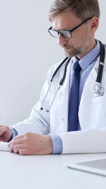 Orta yaşlı bir doktor bir ofis ortamındaki bir panoya reçete yazıyor ve tıbbi kayıtları dolduruyor. Sağlık ve hasta bakımının temel yönlerini gösteriyor. İlaç ve sağlık