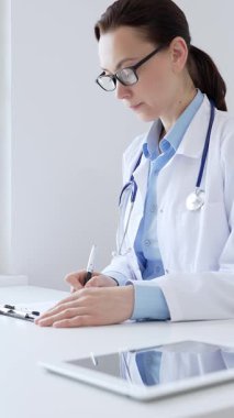 Orta yaşlı kadın doktor, sağlık evraklarını tamamlıyor, dizüstü bilgisayarın yanına yerleştirilmiş bir pano ve steteskop, pencerenin yanında iyi aydınlatılmış klinik çalışma alanı. İlaç ve sağlık hizmetleri.