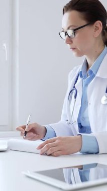Orta yaşlı kadın doktor, sağlık evraklarını tamamlıyor, dizüstü bilgisayarın yanına yerleştirilmiş bir pano ve steteskop, pencerenin yanında iyi aydınlatılmış klinik çalışma alanı. İlaç ve sağlık hizmetleri.