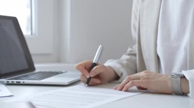 Businesswoman wearing a watch is signing a contract at her office desk, with a laptop and other documents nearby, suggesting a productive workday. Business people concept
