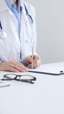 Sağlık evraklarını tamamlayan kadın doktor, dizüstü bilgisayarın yanına yerleştirilmiş bir pano ve cam kenarında iyi aydınlatılmış bir klinik çalışma alanında stetoskop. İlaç ve sağlık hizmetleri kavramı.