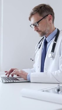 Orta yaşlı doktor özenle bir dizüstü bilgisayarda yazıyor, muhtemelen hasta kayıtlarını güncelliyor ya da araştırma yapıyor, sağlık ve teknolojinin tıbbi ortamdaki kesişimini gösteriyor. İlaç..