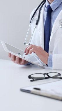 Laboratuvar önlüğü ve steteskop giyen erkek doktor, tıbbi muayenehanede masasında otururken dijital tablet kullanıyor..