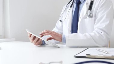 Tıbbi önlük giyen ve steteskop kullanan profesyonel bir doktor, tıp bürosunda masasında otururken dijital tablet kullanıyor, ön planda da tıbbi çizelgesi ve kalemi var. İlaç ve sağlık hizmetleri.