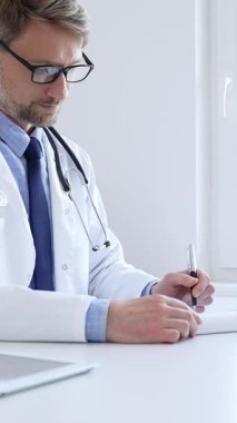 Orta yaşlı bir doktor bir ofis ortamındaki bir panoya reçete yazıyor ve tıbbi kayıtları dolduruyor. Sağlık ve hasta bakımının temel yönlerini gösteriyor. İlaç ve sağlık