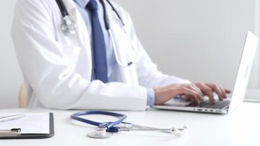 Mavi steteskop, klinikteki masada otururken dizüstü bilgisayarda yazan doktor profesyonelin yanındaki masada yatıyor. Tıp ve sağlık hizmetleri kavramı.