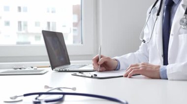 Erkek doktor reçete yazıyor ve bir ofis ortamındaki bir panoya tıbbi kayıtları dolduruyor. Sağlık ve hasta bakımının temel yönlerini gösteriyor. İlaç ve sağlık hizmetleri.