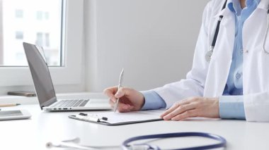 Sağlık evraklarını tamamlayan kadın doktor, dizüstü bilgisayarın yanına yerleştirilmiş bir pano ve cam kenarında iyi aydınlatılmış bir klinik çalışma alanında stetoskop. İlaç ve sağlık hizmetleri kavramı.