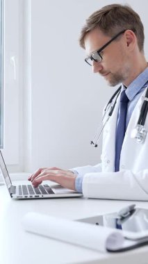 Orta yaşlı doktor özenle bir dizüstü bilgisayarda yazıyor, muhtemelen hasta kayıtlarını güncelliyor ya da araştırma yapıyor, sağlık ve teknolojinin tıbbi ortamdaki kesişimini gösteriyor. İlaç..