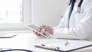 Tıbbi önlük giyen ve steteskop kullanan profesyonel bir doktor, tıp bürosunda masasında otururken dijital tablet kullanıyor, ön planda da tıbbi çizelgesi ve kalemi var. İlaç ve sağlık hizmetleri.