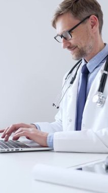 Orta yaşlı doktor özenle bir dizüstü bilgisayarda yazıyor, muhtemelen hasta kayıtlarını güncelliyor ya da araştırma yapıyor, sağlık ve teknolojinin tıbbi ortamdaki kesişimini gösteriyor. İlaç..