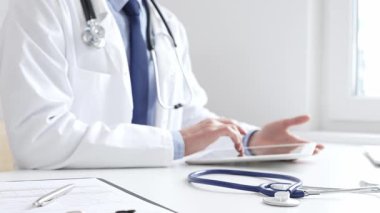 Klinikte dijital tablet kullanan doktor adamın yanındaki masada mavi steteskop duruyor. Tıp ve sağlık hizmetleri kavramı