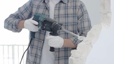 İnşaat işçisi, beyaz duvarı döner çekiçli matkapla yıkıyor, gri kareli gömlek ve koruyucu beyaz eldivenler giyiyor, yenileme projesi sırasında toz üretiyor, yakın plan görüntüsü var..