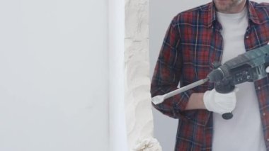 İnşaat işçisi, beyaz duvarı döner çekiç matkabıyla yıkıyor, kırmızı kareli gömlek ve koruyucu beyaz eldivenler giyiyor, yenileme projesi sırasında toz üretiyor, yakın çekim görüntüsü sağlıyor..
