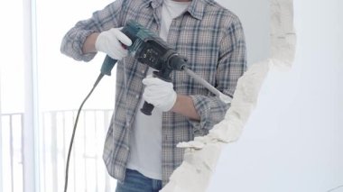 İnşaat işçisi, beyaz duvarı döner çekiçli matkapla yıkıyor, gri kareli gömlek ve koruyucu beyaz eldivenler giyiyor, yenileme projesi sırasında toz üretiyor, yakın plan görüntüsü var..