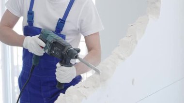 İnşaat işçisi, beyaz duvarı döner çekiç matkabıyla yıkıyor, mavi tulum ve koruyucu beyaz eldivenler giyiyor, yenileme projesi sırasında toz üretiyor, yakın çekim görüntüsü sağlıyor..