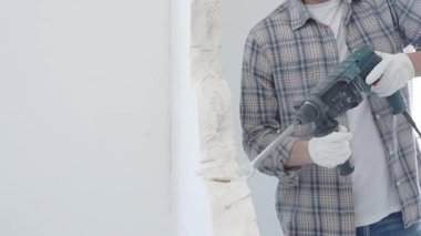 İnşaat işçisi, beyaz duvarı döner çekiçli matkapla yıkıyor, gri kareli gömlek ve koruyucu beyaz eldivenler giyiyor, yenileme projesi sırasında toz üretiyor, yakın plan görüntüsü var..
