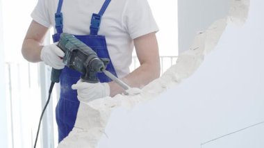 İnşaat işçisi, beyaz duvarı döner çekiç matkabıyla yıkıyor, mavi tulum ve koruyucu beyaz eldivenler giyiyor, yenileme projesi sırasında toz üretiyor, yakın çekim görüntüsü sağlıyor..