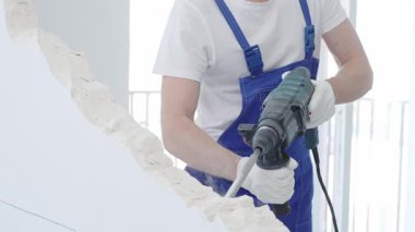 İnşaat işçisi, beyaz duvarı döner çekiç matkabıyla yıkıyor, mavi tulum ve koruyucu beyaz eldivenler giyiyor, yenileme projesi sırasında toz üretiyor, yakın çekim görüntüsü sağlıyor..