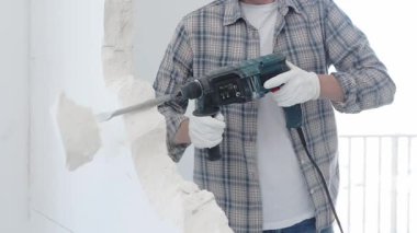 İnşaat işçisi, beyaz duvarı döner çekiçli matkapla yıkıyor, gri kareli gömlek ve koruyucu beyaz eldivenler giyiyor, yenileme projesi sırasında toz üretiyor, yakın plan görüntüsü var..