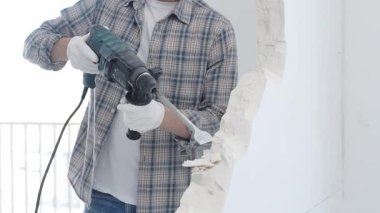 İnşaat işçisi, beyaz duvarı döner çekiçli matkapla yıkıyor, gri kareli gömlek ve koruyucu beyaz eldivenler giyiyor, yenileme projesi sırasında toz üretiyor, yakın plan görüntüsü var..