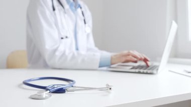 Mavi steteskopun kapatılması, dizüstü bilgisayarda çalışan kadın doktorun yanındaki masada yatıyor ve sağlık hizmetleri teşhisi ve hasta belgelerini dönüştüren dijital araçları vurguluyor. Tıp konsepti.