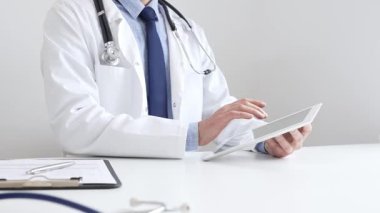 Tıbbi önlük giyen ve steteskop kullanan profesyonel bir doktor, tıp bürosunda masasında otururken dijital tablet kullanıyor, ön planda da tıbbi çizelgesi ve kalemi var. İlaç ve sağlık hizmetleri.