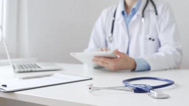 Mavi steteskopun yakın çekimleri kadın doktorun yanında duruyor. Tabletteki hasta verilerini gözden geçiriyor ve çağdaş tıbbi ortamdaki dijital sağlık hizmetleri akışını temsil ediyor..