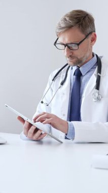 Laboratuvar önlüğü ve steteskop giyen orta yaşlı doktor, tıp bürosunda masasında otururken dijital tablet kullanıyor, ön planda da da tıbbi çizelge ve kalem var. Tıp konsepti.