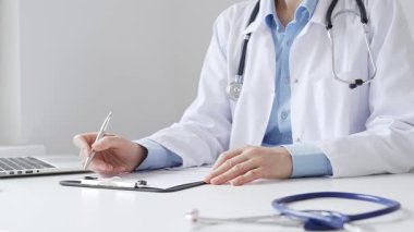 Sağlık evraklarını tamamlayan kadın doktor, dizüstü bilgisayarın yanına yerleştirilmiş bir pano ve cam kenarında iyi aydınlatılmış bir klinik çalışma alanında stetoskop. İlaç ve sağlık hizmetleri kavramı.