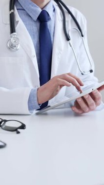 Laboratuvar önlüğü ve steteskop giyen erkek doktor, tıbbi muayenehanede masasında otururken dijital tablet kullanıyor..