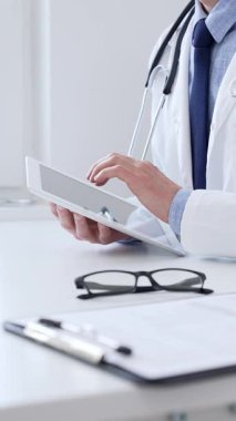 Laboratuvar önlüğü ve steteskop giyen erkek doktor, tıbbi muayenehanede masasında otururken dijital tablet kullanıyor..