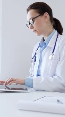 Orta yaşlı doktor kadın laboratuvar önlüğü ve steteskop giyiyordu. Dijital tablet kullanıyordu. Bilgisayarı ve tıbbi kayıtları, modern sağlık teknolojisini sergiliyordu. İlaç ve sağlık
