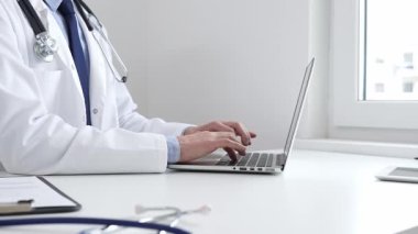 Doktor özenle bir dizüstü bilgisayarda yazıyor, muhtemelen hasta kayıtlarını güncelliyor veya araştırma yapıyor, modern bir tıbbi ortamda sağlık ve teknolojinin kesişimini gösteriyor.