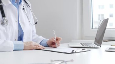 Sağlık evraklarını tamamlayan kadın doktor, dizüstü bilgisayarın yanına yerleştirilmiş bir pano ve cam kenarında iyi aydınlatılmış bir klinik çalışma alanında stetoskop. İlaç ve sağlık hizmetleri kavramı.