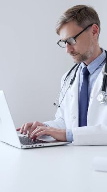 Orta yaşlı doktor özenle bir dizüstü bilgisayarda yazıyor, muhtemelen hasta kayıtlarını güncelliyor ya da araştırma yapıyor, sağlık ve teknolojinin tıbbi ortamdaki kesişimini gösteriyor. İlaç..