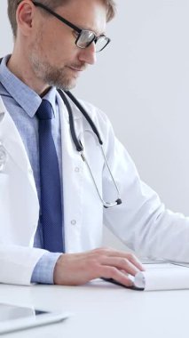 Orta yaşlı bir doktor bir ofis ortamındaki bir panoya reçete yazıyor ve tıbbi kayıtları dolduruyor. Sağlık ve hasta bakımının temel yönlerini gösteriyor. İlaç ve sağlık