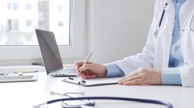 Sağlık evraklarını tamamlayan kadın doktor, dizüstü bilgisayarın yanına yerleştirilmiş bir pano ve cam kenarında iyi aydınlatılmış bir klinik çalışma alanında stetoskop. İlaç ve sağlık hizmetleri kavramı.