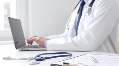 Mavi steteskop, klinikteki masada otururken dizüstü bilgisayarda yazan doktor profesyonelin yanındaki masada yatıyor. Tıp ve sağlık hizmetleri kavramı.