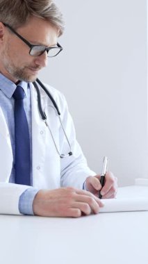 Orta yaşlı bir doktor bir ofis ortamındaki bir panoya reçete yazıyor ve tıbbi kayıtları dolduruyor. Sağlık ve hasta bakımının temel yönlerini gösteriyor. İlaç ve sağlık