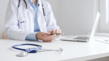 Mavi steteskopun yakın çekimleri, iyi aydınlatılmış klinik çalışma alanındaki dijital sağlık hizmetlerini temsil eden akıllı telefonu kontrol eden kadın doktorun yanındaki masada yatıyor. Tıp konsepti.