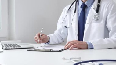 Erkek doktor reçete yazıyor ve bir ofis ortamındaki bir panoya tıbbi kayıtları dolduruyor. Sağlık ve hasta bakımının temel yönlerini gösteriyor. İlaç ve sağlık hizmetleri.