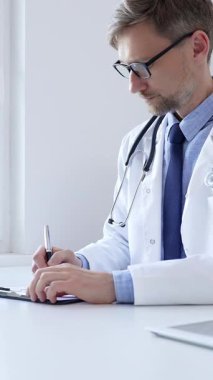 Orta yaşlı bir doktor bir ofis ortamındaki bir panoya reçete yazıyor ve tıbbi kayıtları dolduruyor. Sağlık ve hasta bakımının temel yönlerini gösteriyor. İlaç ve sağlık