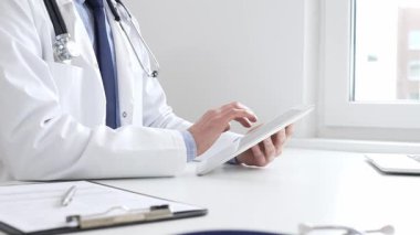 Laboratuvar önlüğü ve steteskop giyen erkek doktor, tıbbi muayenehanede masasında otururken dijital tablet kullanıyor..